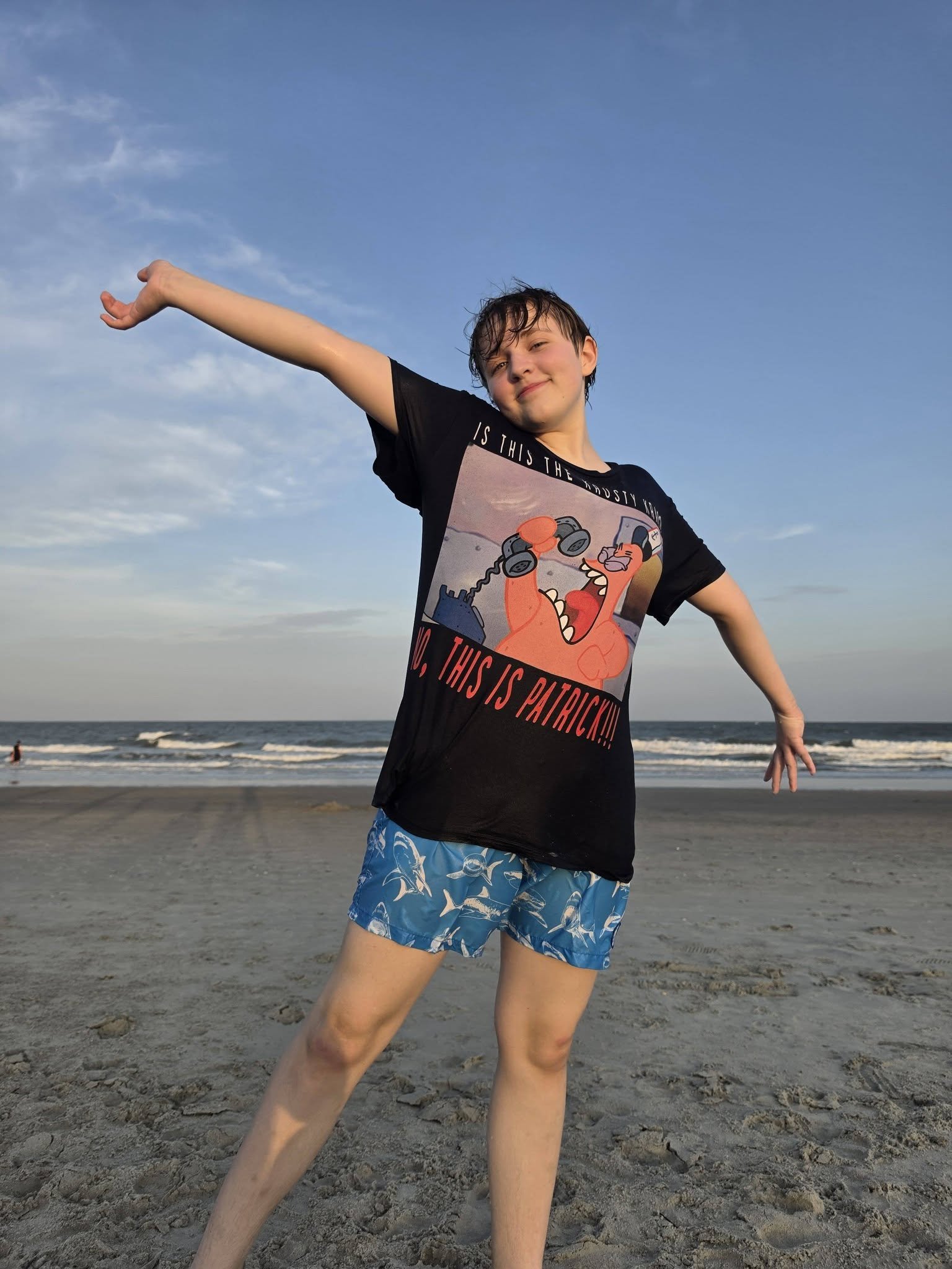 A boy standing on a sandy beach with his arms outstretched, wearing a black t-shirt with cartoon characters and blue shorts with white patterns, smiling with the ocean and sky in the background.