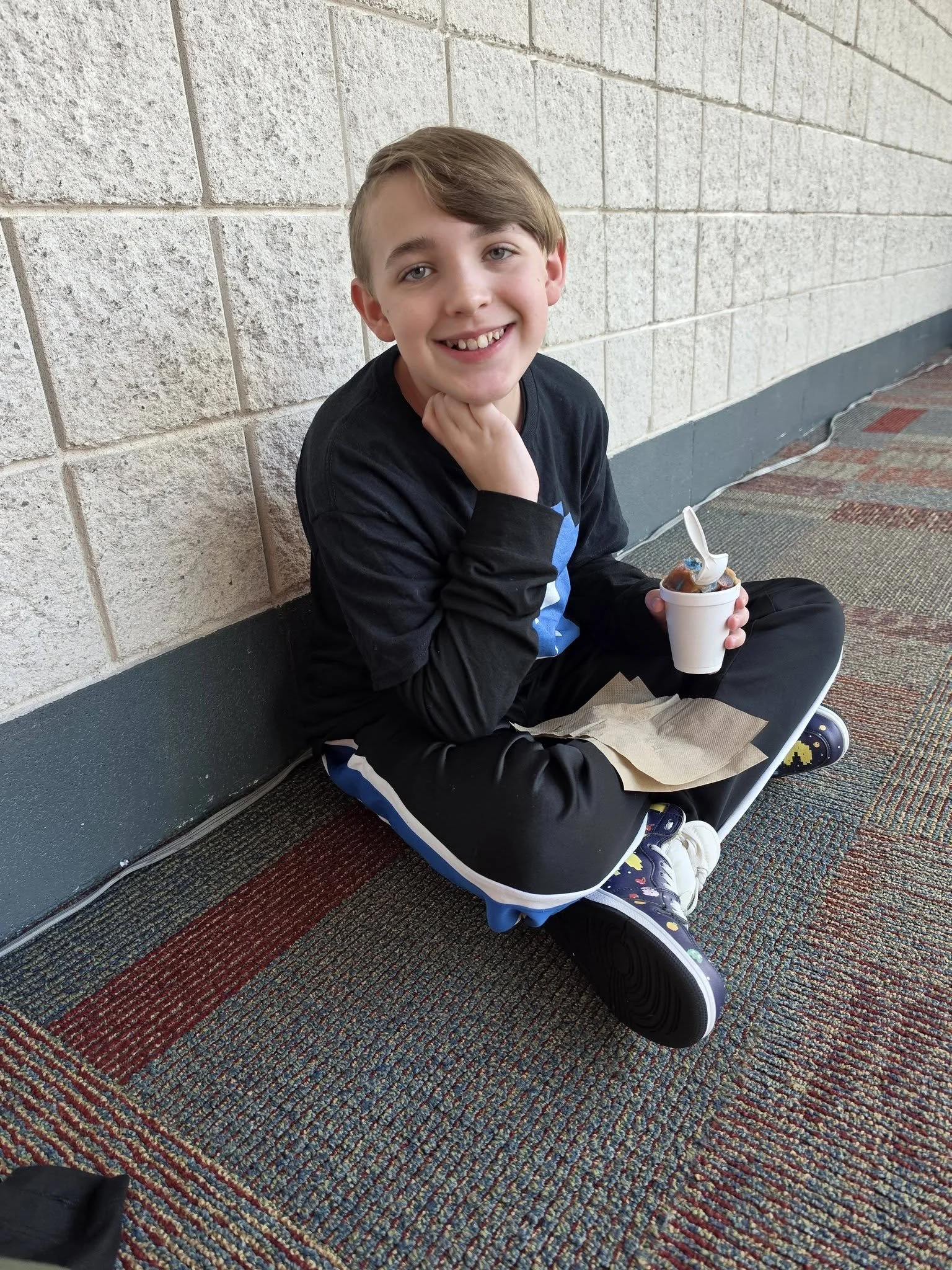 A smiling boy sitting on the floor against a beige brick wall, holding a cup of ice cream with a plastic spoon, wearing a black and blue shirt and patterned sneakers.