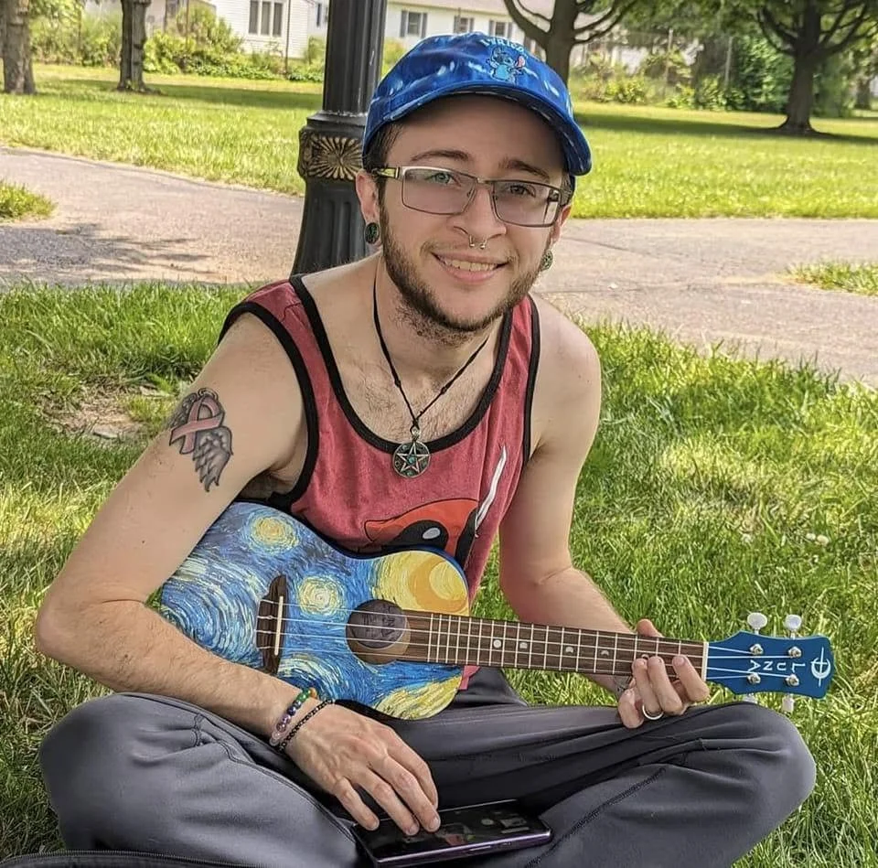 A young man with glasses and tattoos sits on the grass in a park, smiling and holding a small, painted ukulele. He is wearing a blue cap, a sleeveless tank top, and has a phone in his lap.