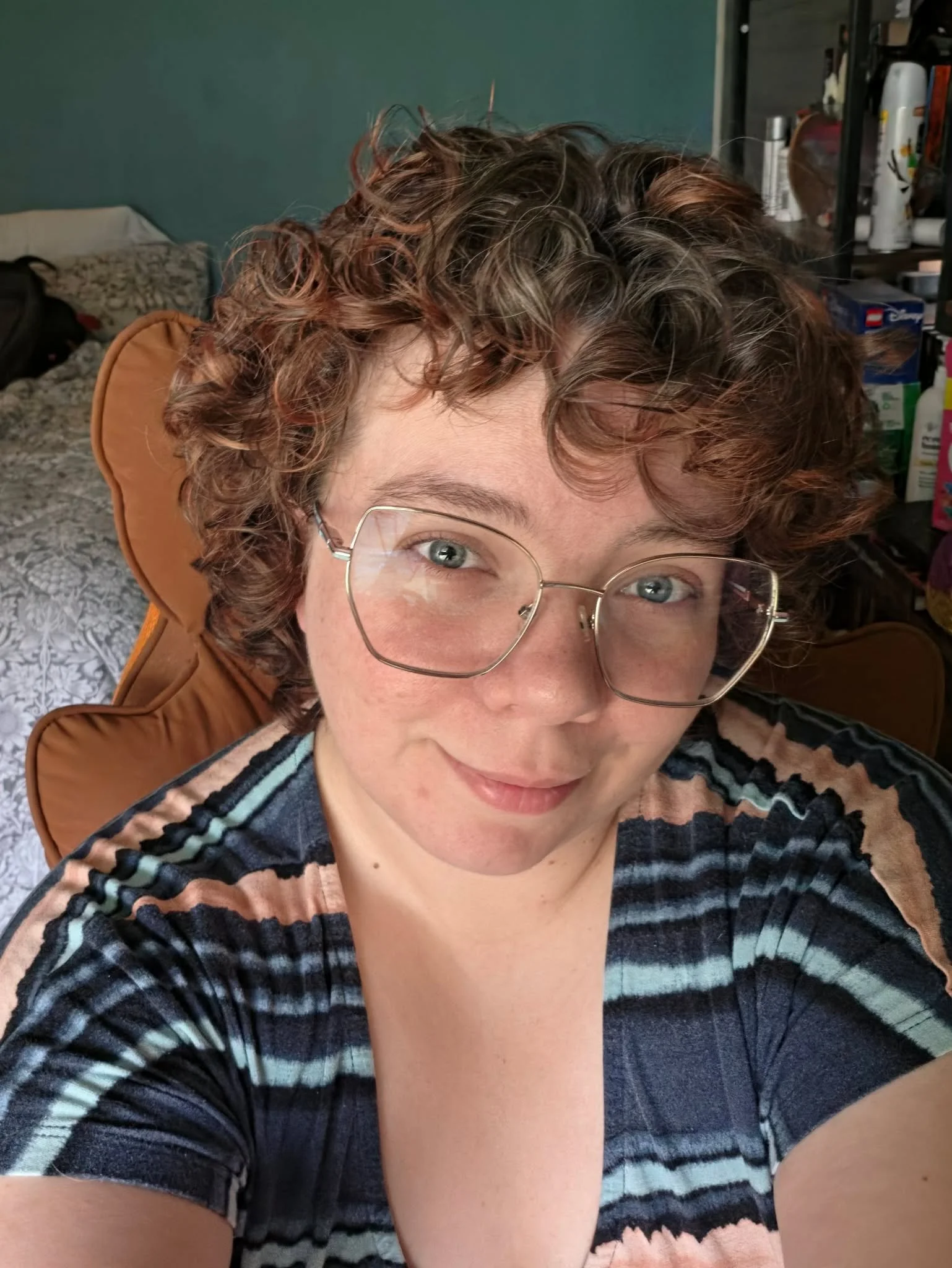 A woman with curly short hair wearing glasses and a striped shirt, looking into the camera. Inside a room with a bed and shelves in the background.