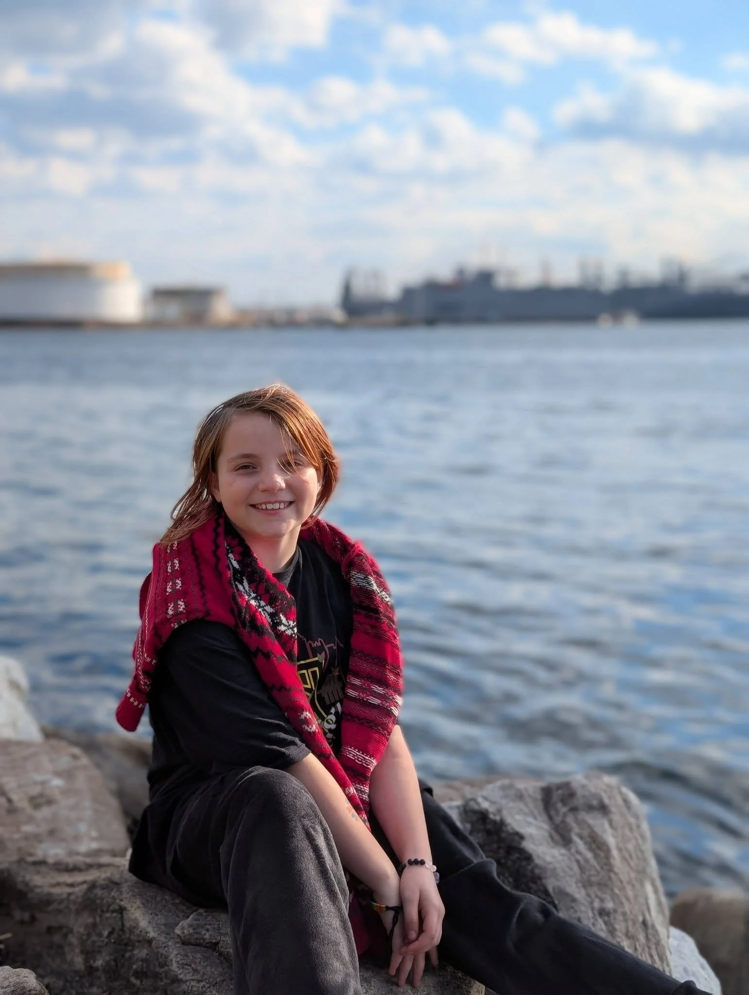 A young woman with shoulder-length red hair sitting on rocks near a body of water, smiling at the camera, with ships and a blue sky with clouds in the background.