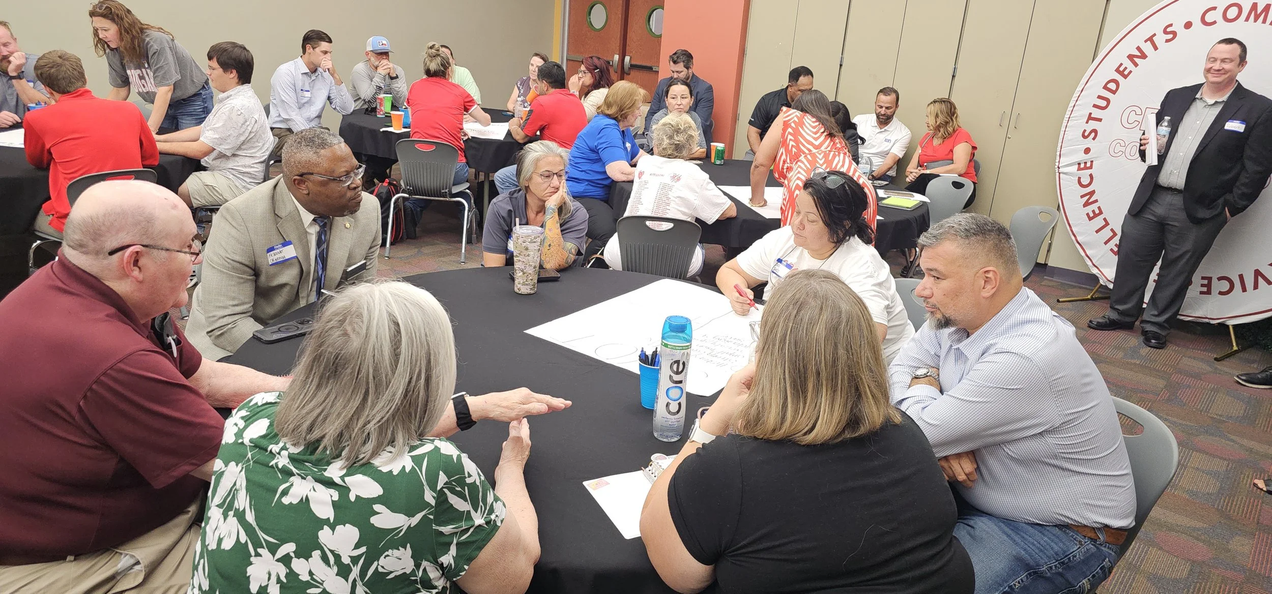 A diverse group of people, including men and women of different ages and ethnicities, gathered around tables in a conference or workshop setting. Some are engaged in discussion, writing, or listening, while others are observing. The room has beige an