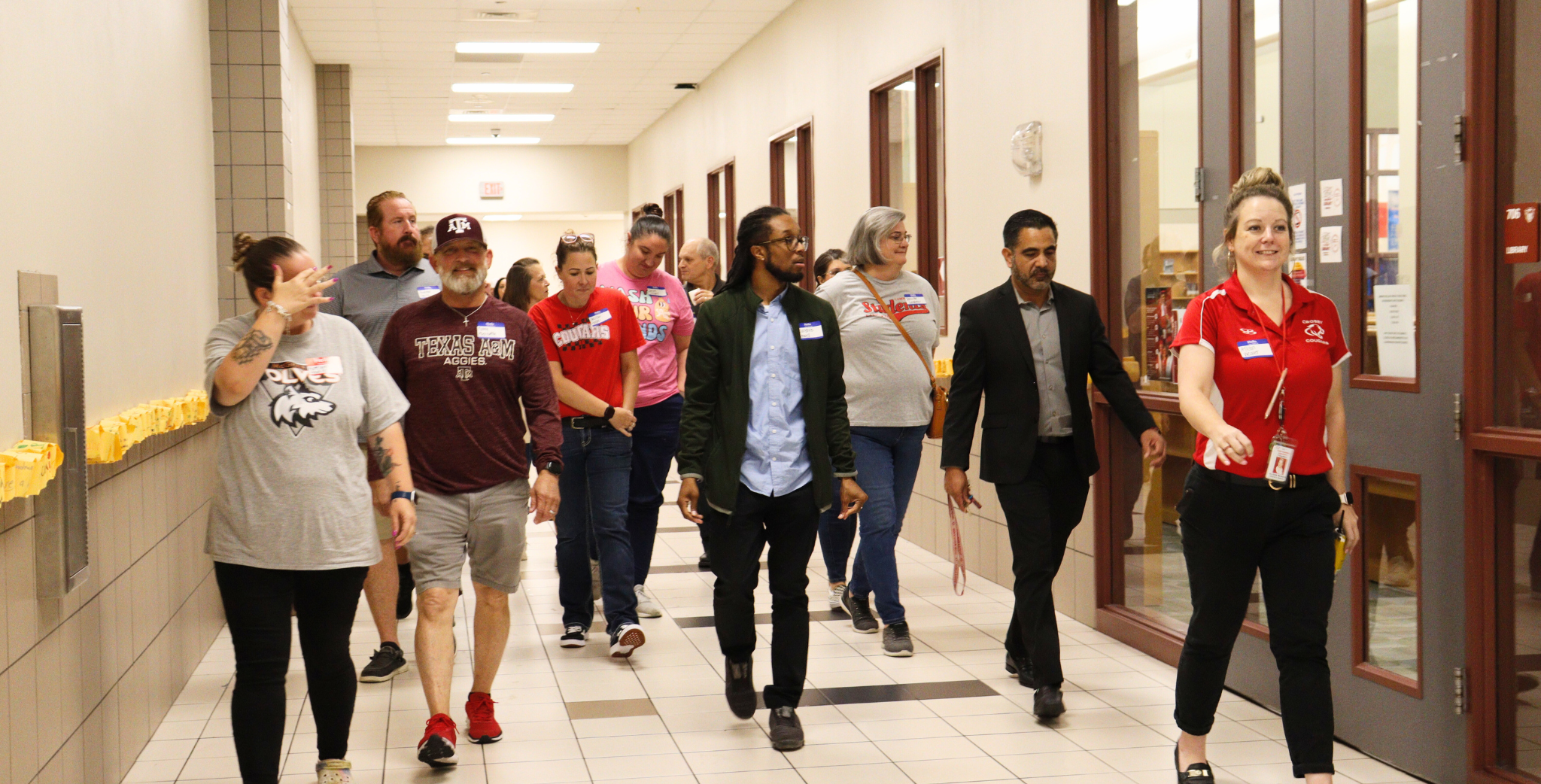 People walking down a school hallway, some wearing college or university apparel, with a woman in a red shirt leading, and others engaging in conversations.