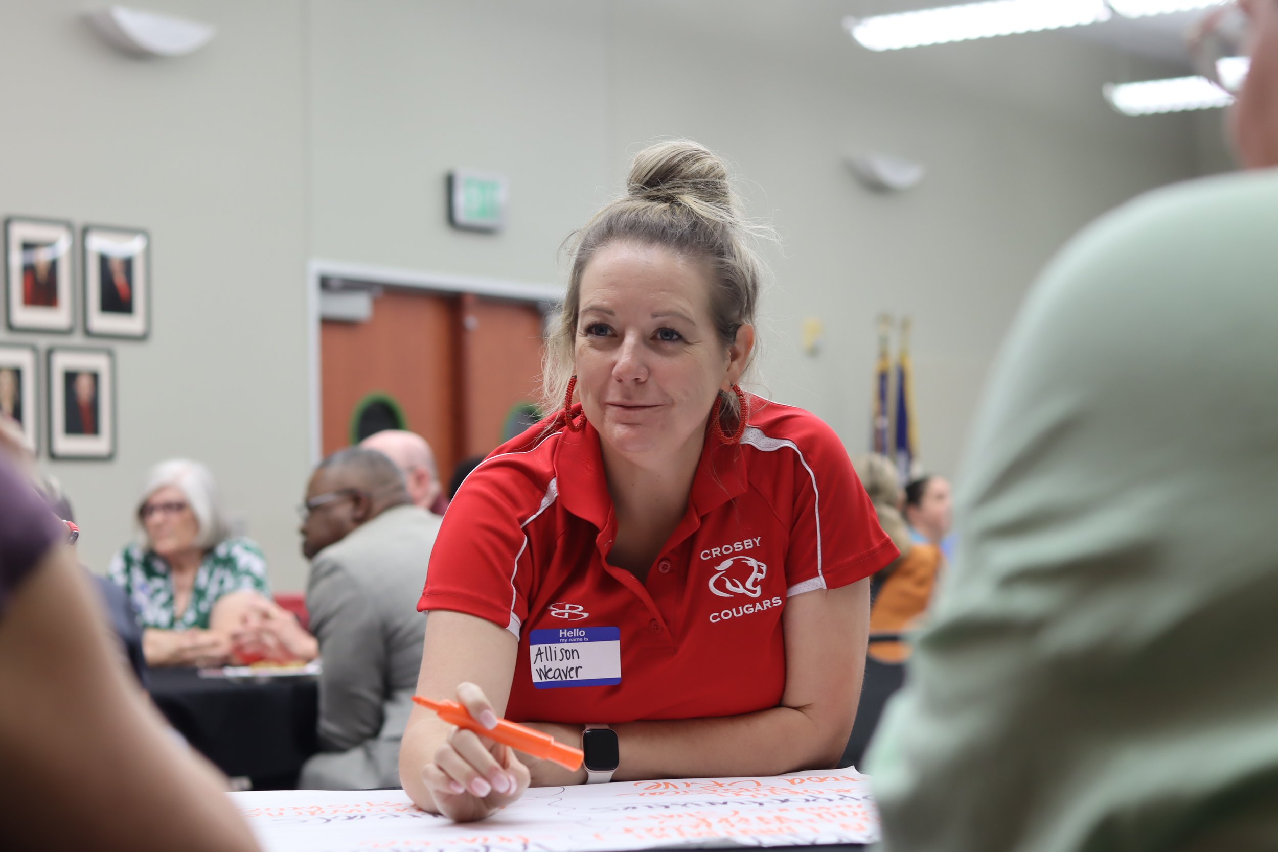 A woman wearing a red shirt with a Crosby Cougar logo and a name tag that reads "Allison Weaver" is sitting at a table during an indoor group event. She has her hair in a top knot and is holding an orange marker, engaged in conversation or activity with several other people around her.