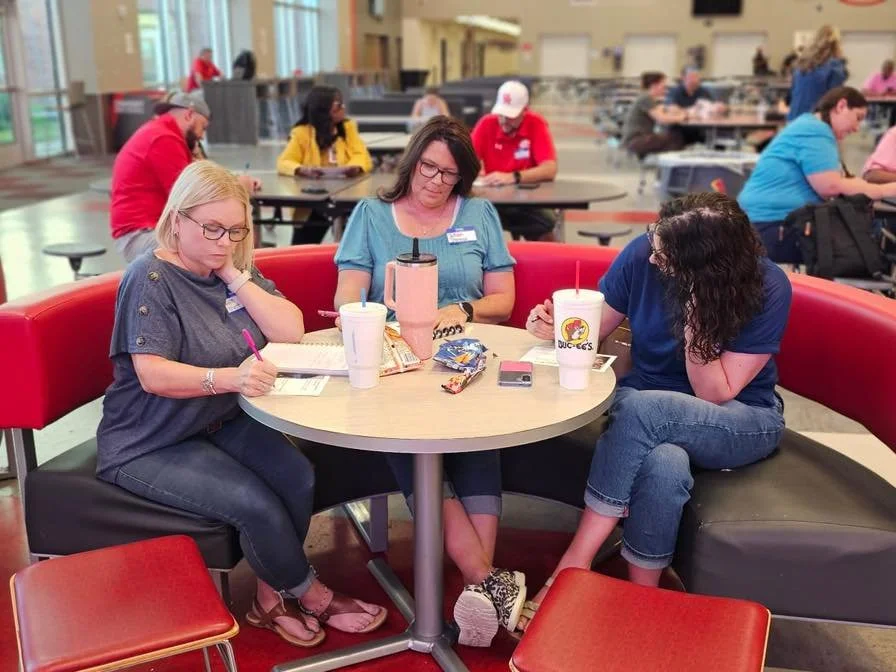Three women sitting at a round table with fast food drinks in an airport terminal, engaged in a game or activity.