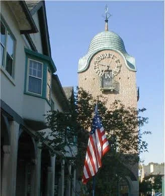 A image of the Historic Market Square with a tall clock tower with a green domed roof, a weather vane on top, and Roman numerals on the clock face, with an American flag in front of a building under a clear blue sky in Lake Forest, IL.