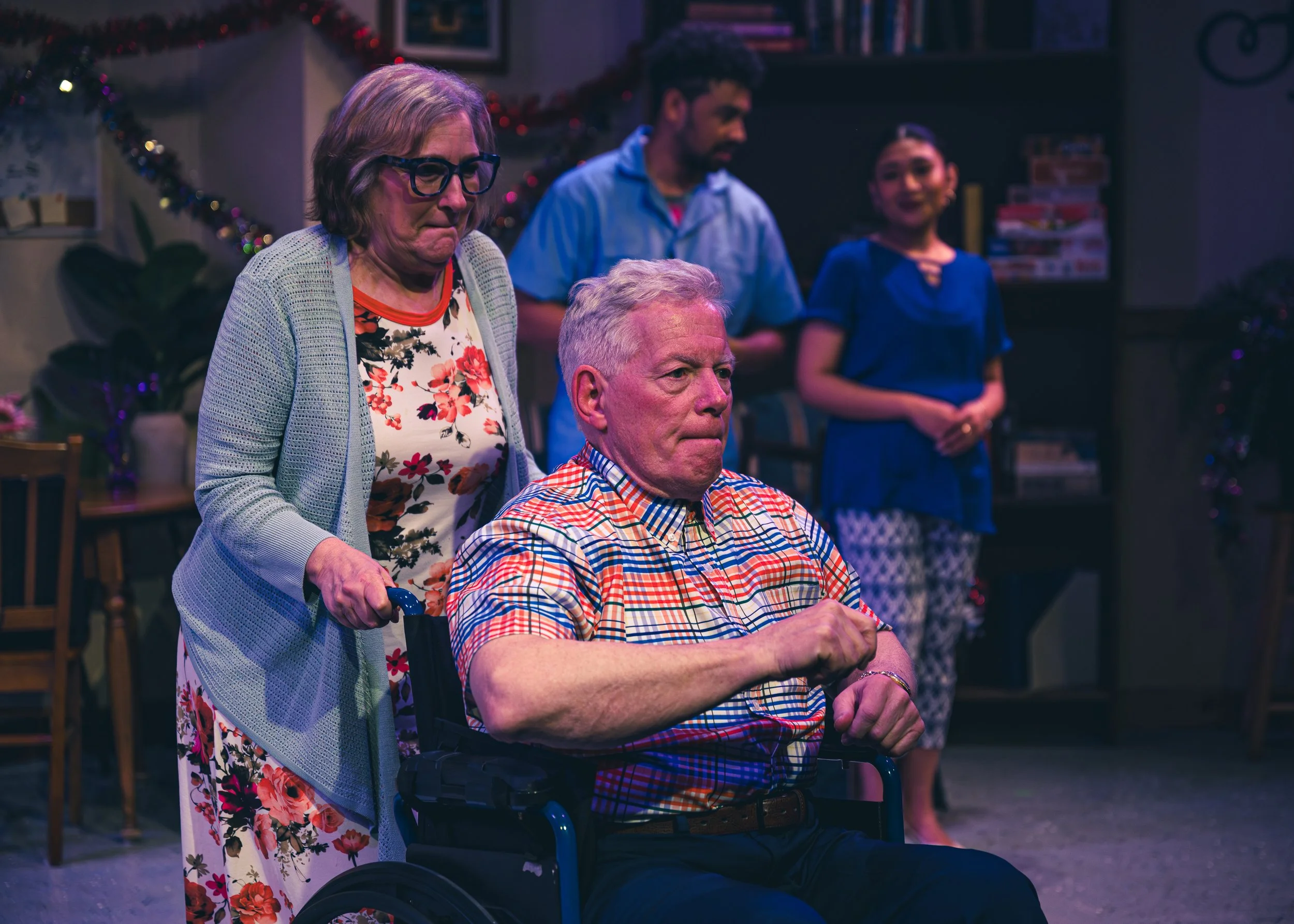 A woman with grey hair pushes a man with grey hair in a wheelchar. A young man and a young woman appear in the background. A scene from Citadel's production of The Jukebox.