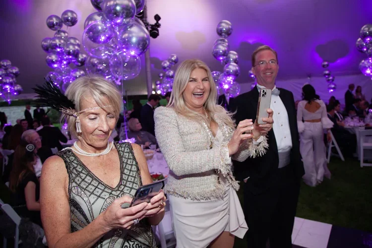 Three people, two women and one man, at a celebration or event in a decorated tent with purple lighting and balloon decorations. They are smiling and looking at their phones, with other guests in the background.