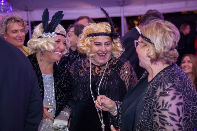 Group of women dressed in vintage 1920s-style flapper costumes and accessories, smiling and talking at a lively party or event.