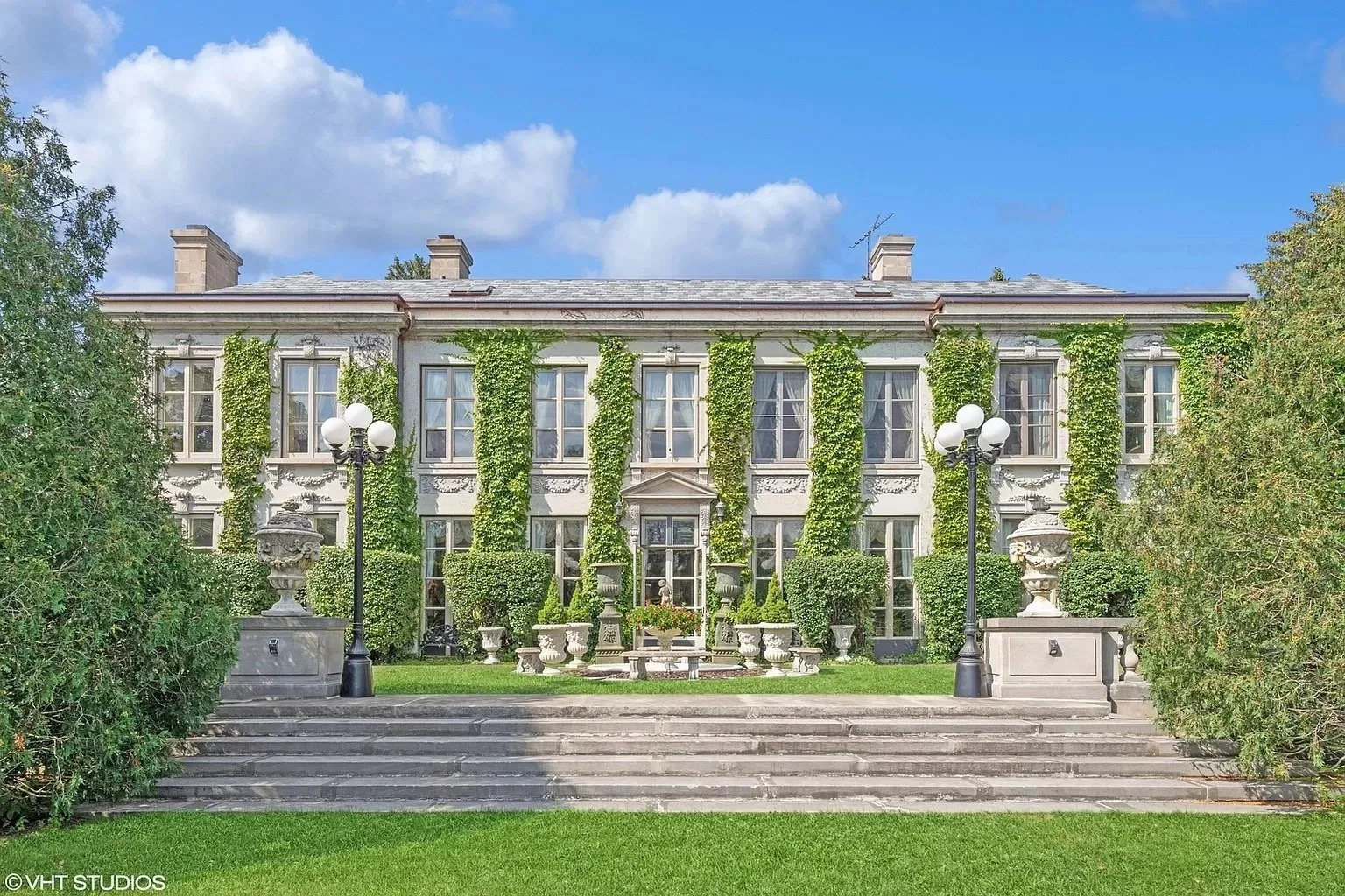 Large white historic mansion called the Adler Estate with tall windows covered in green vines, stone steps leading to the entrance, decorative urns, benches, and street lamps in front, under a partly cloudy blue sky.