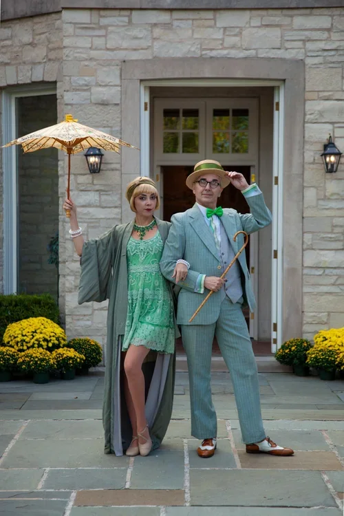 Two elegantly dressed individuals, a woman and a man, standing outside a stone building with open doors, surrounded by yellow chrysanthemums. The woman holds a white parasol and wears a green dress with a matching cape, while the man wears a light bl