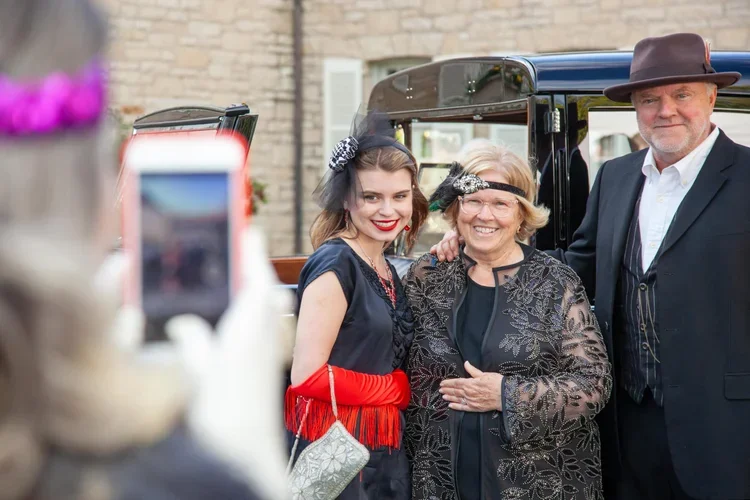 Three people dressed in vintage clothing posing for a photo outdoors, with a brick building in the background. The person on the left is a young woman with brown hair, wearing a dark dress, red gloves, and a black fascinator. The woman in the middle 