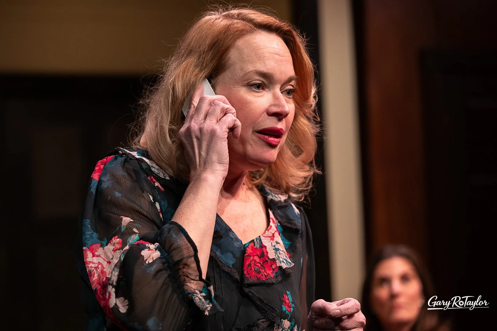 A woman with red hair, wearing a black floral blouse, is talking on a cellphone during a meeting or event. A woman with dark hair is visible in the background. From a Citadel Theatre production.