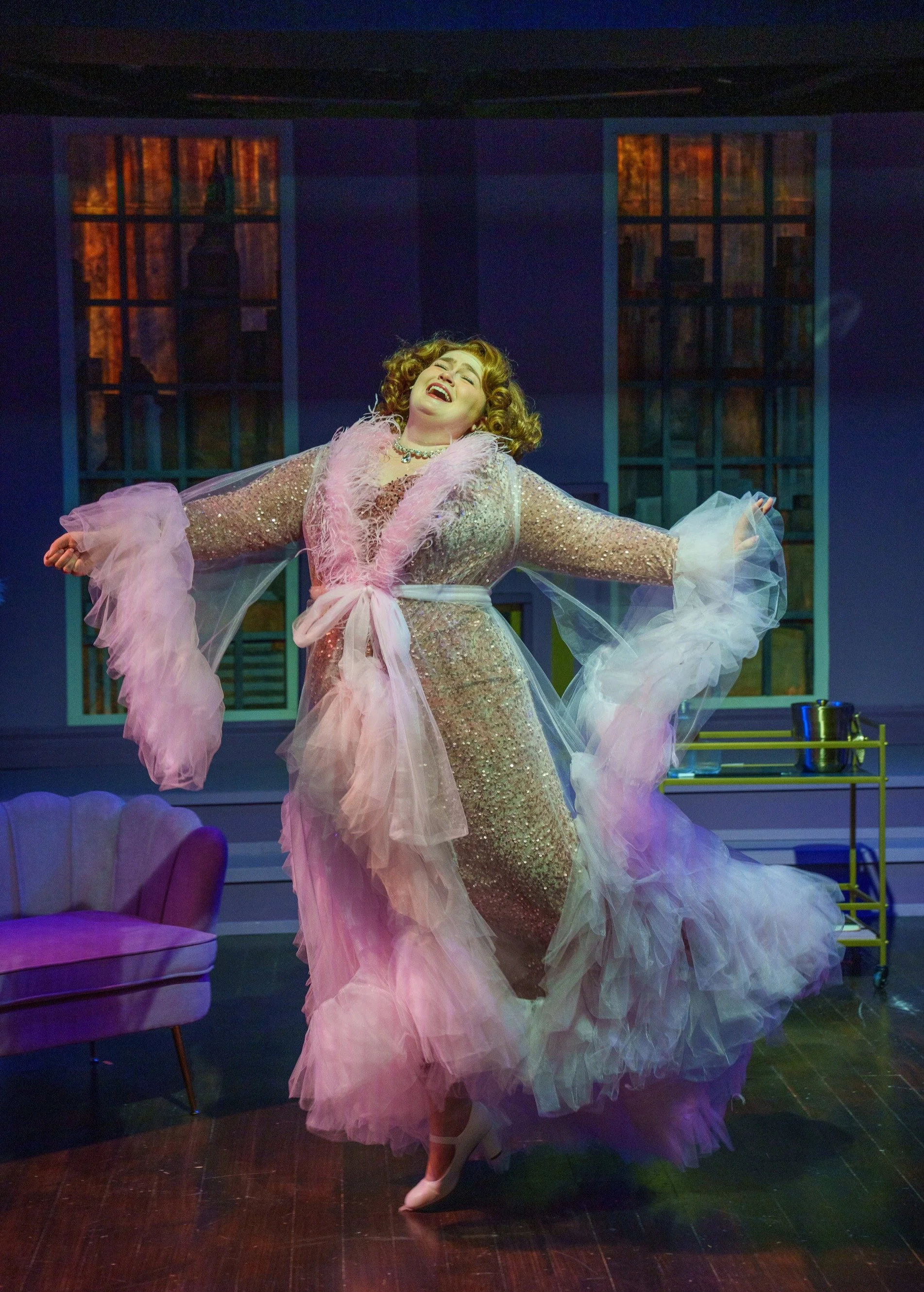 A woman performing on stage, dressed in a glamorous, long pink and gold gown with ruffled tulle details, singing with her arms outstretched in front of a blue set with large windows. She has curly blonde hair and appears to be in the middle of a song