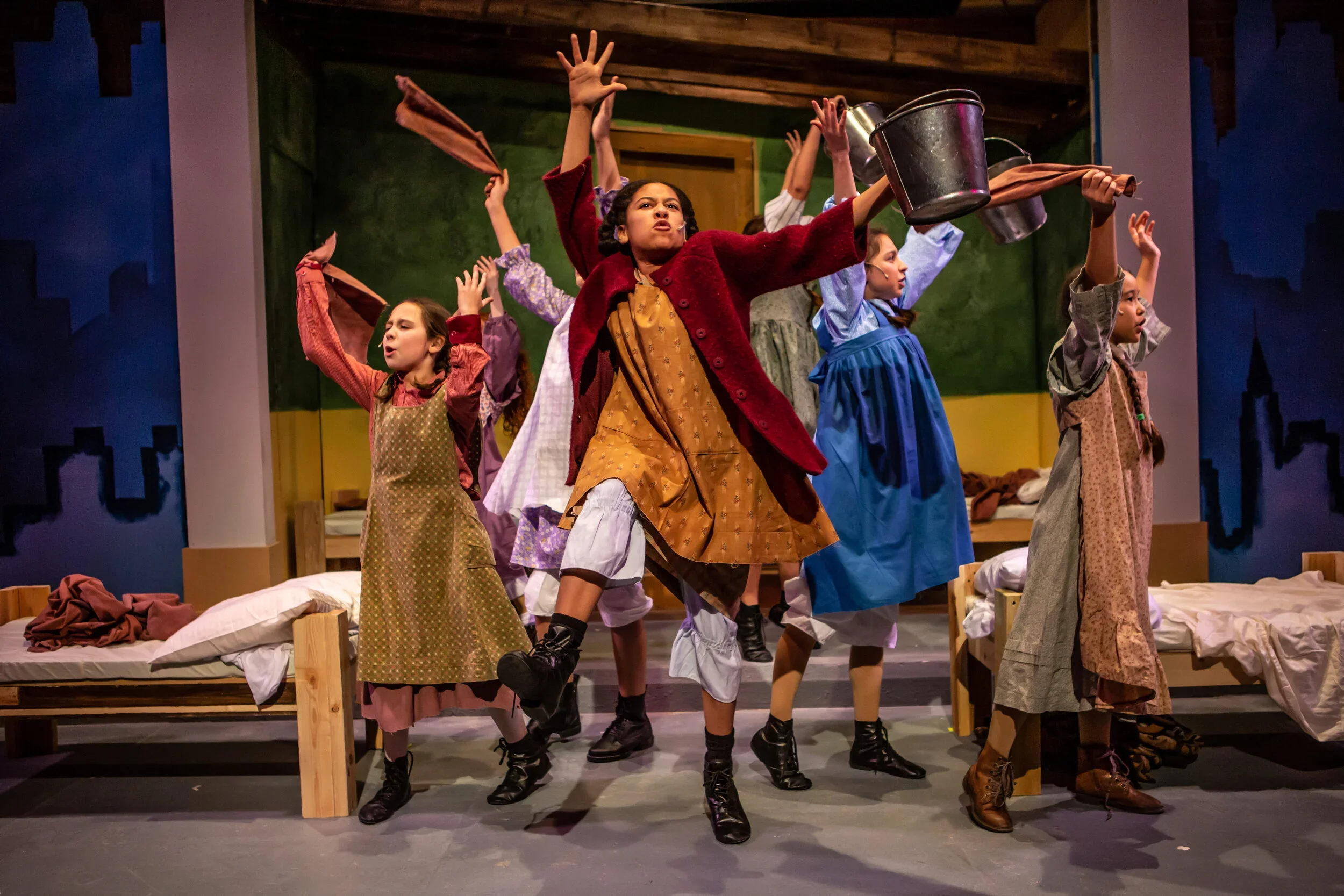 Children performing It's a Hard Knock Life in Citadel's production of Annie on a stage in a play, dressed in old-fashioned costumes, with some raising their hands and holding buckets, set in a rustic bedroom scene.