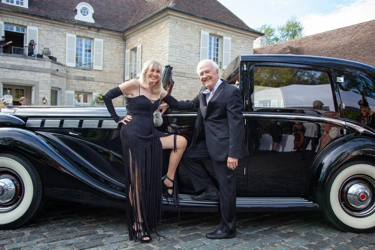 A woman in a black evening gown and black gloves, sitting on the door of a vintage black car, smiling next to a man in a black suit and bow tie, posing in front of a large stone house.