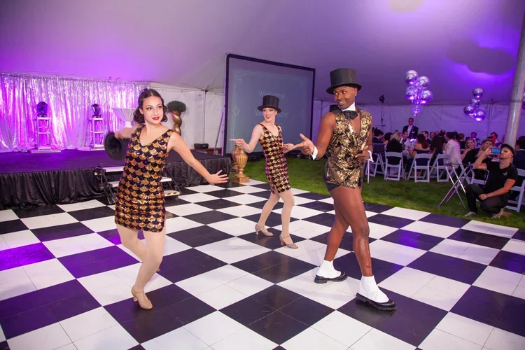 Three performers in vintage gold and black costumes dancing on a black and white checkered floor at an event. Two women wear matching dresses and hats, while a man in a gold vest, shorts, top hat, and white socks with black shoes participates. Audien
