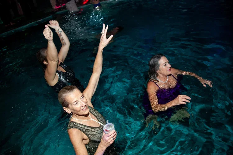 Women enjoying swimming in a pool at a party, dancing and holding drinks.