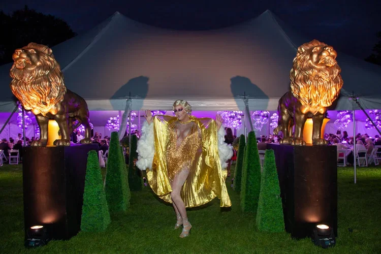 A person dressed in a gold, sequined outfit with a cape, walking through an outdoor event area illuminated with purple lighting, flanked by large lion statues on pedestals, with a large tent in the background.