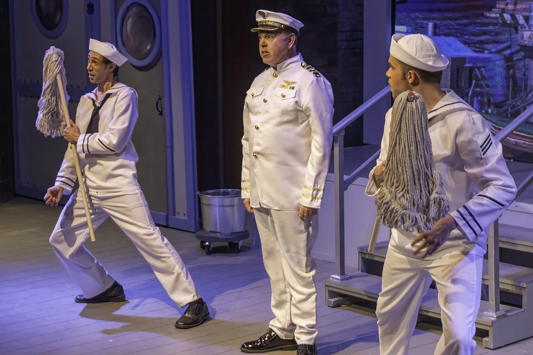 From Citadel's production of Dames at Sea. Three men dressed as sailors in white uniforms with navy hats and mops, standing indoors on a stage with a background of laundry machines and a staircase.
