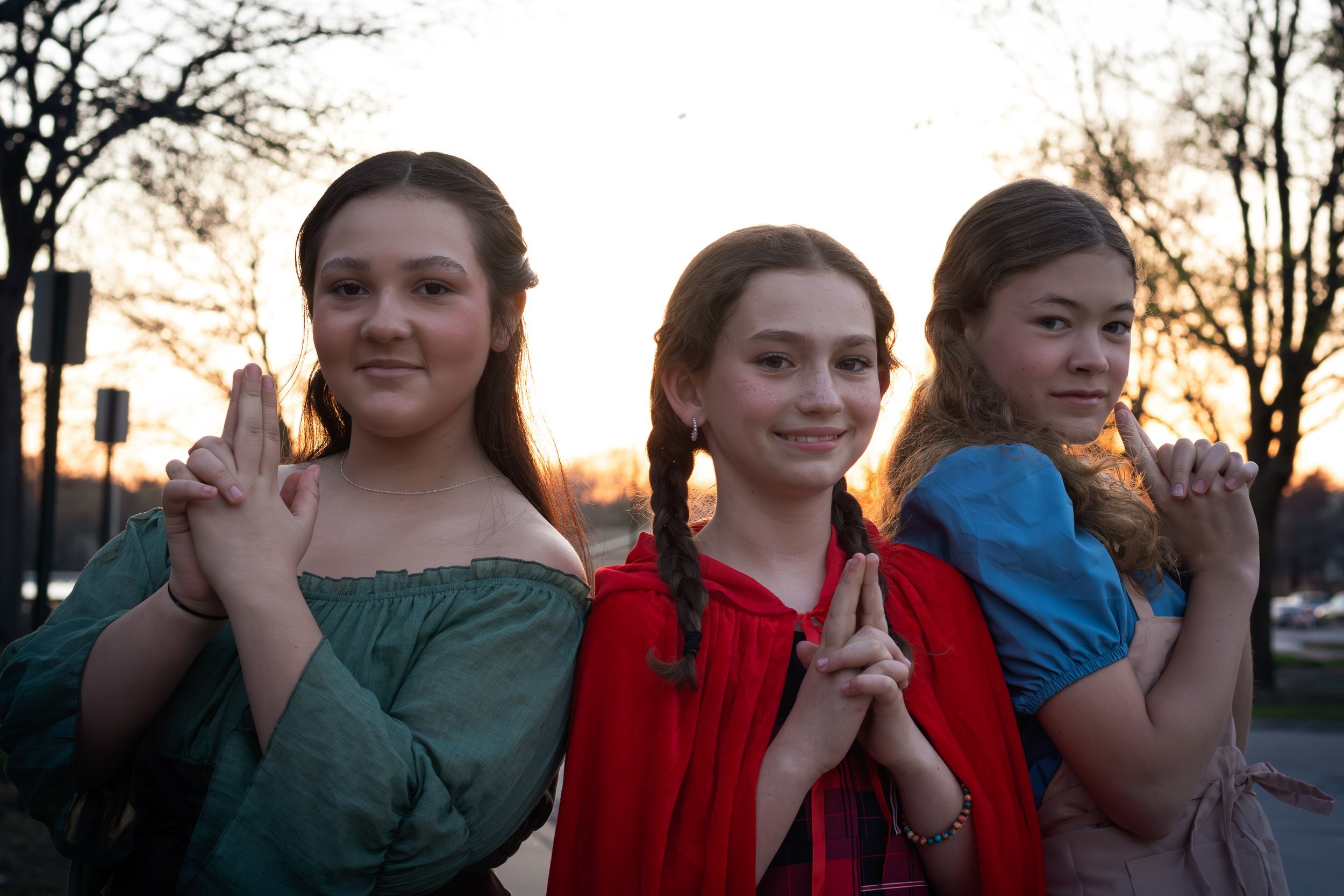 Three young girls standing outdoors at sunset, with trees in the background, making peace signs with their hands.