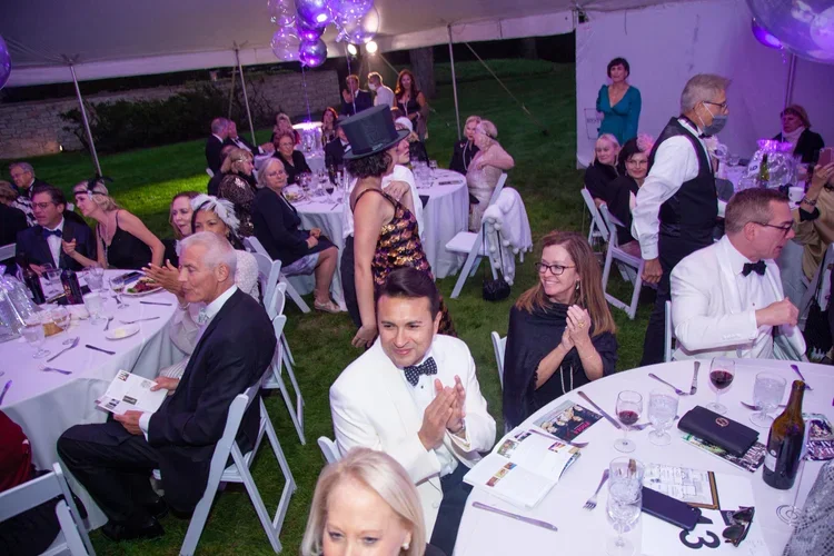 Guests attending a formal outdoor event under a white tent, seated at decorated tables with wine glasses and menus, dressed in tuxedos and evening gowns, some clapping, and a woman in a shiny dress and top hat standing among them.