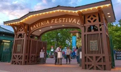 Entrance archway for Ravinia Festival with wooden structure and lights, with people gathering underneath on a walkway surrounded by trees.