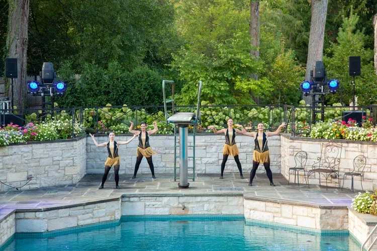 Four dancers in black and gold costumes performing poolside on an outdoor stage with trees and flowers in the background.
