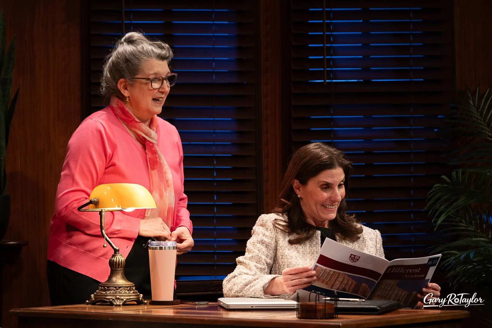 Citadel's  Admissions with two women in office setting, one standing and one sitting. The standing woman is wearing a pink blouse and scarf, and the seated woman is in a light-colored blazer, with a desk lamp, tumbler, and laptop on the desk.