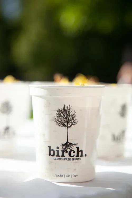 White cup with a black leafless tree logo and the words "birch. GLUTEN FREE SPIRITS" along with vodka, gin, and rum listed below, with background of blurred cups and greenery.