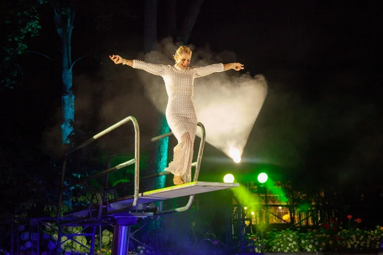 A woman dressed in a white dress is standing on a platform with her arms outstretched, with stage lights and smoke creating a dramatic background.