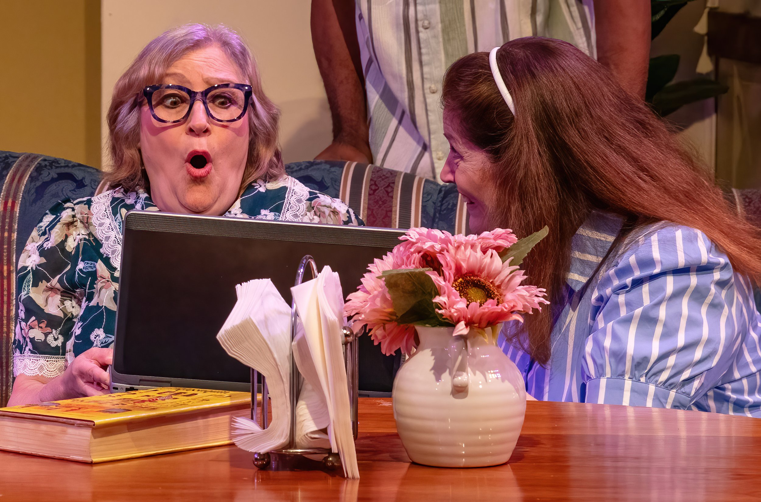 A woman with glasses and a floral shirt looks surprised at a laptop in a cozy room. A young woman with red hair, a white headband, and a striped shirt leans in smiling. A man in a striped shirt stands behind them. The table has a vase of pink flowers, napkins, and a menu.