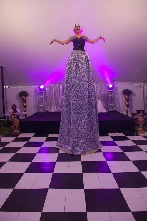 A performer on stilts wearing a long patterned dress, a black and white top, sunglasses, large earrings, and a wig, standing on a checkered dance floor with purple stage lighting and curtains in the background.