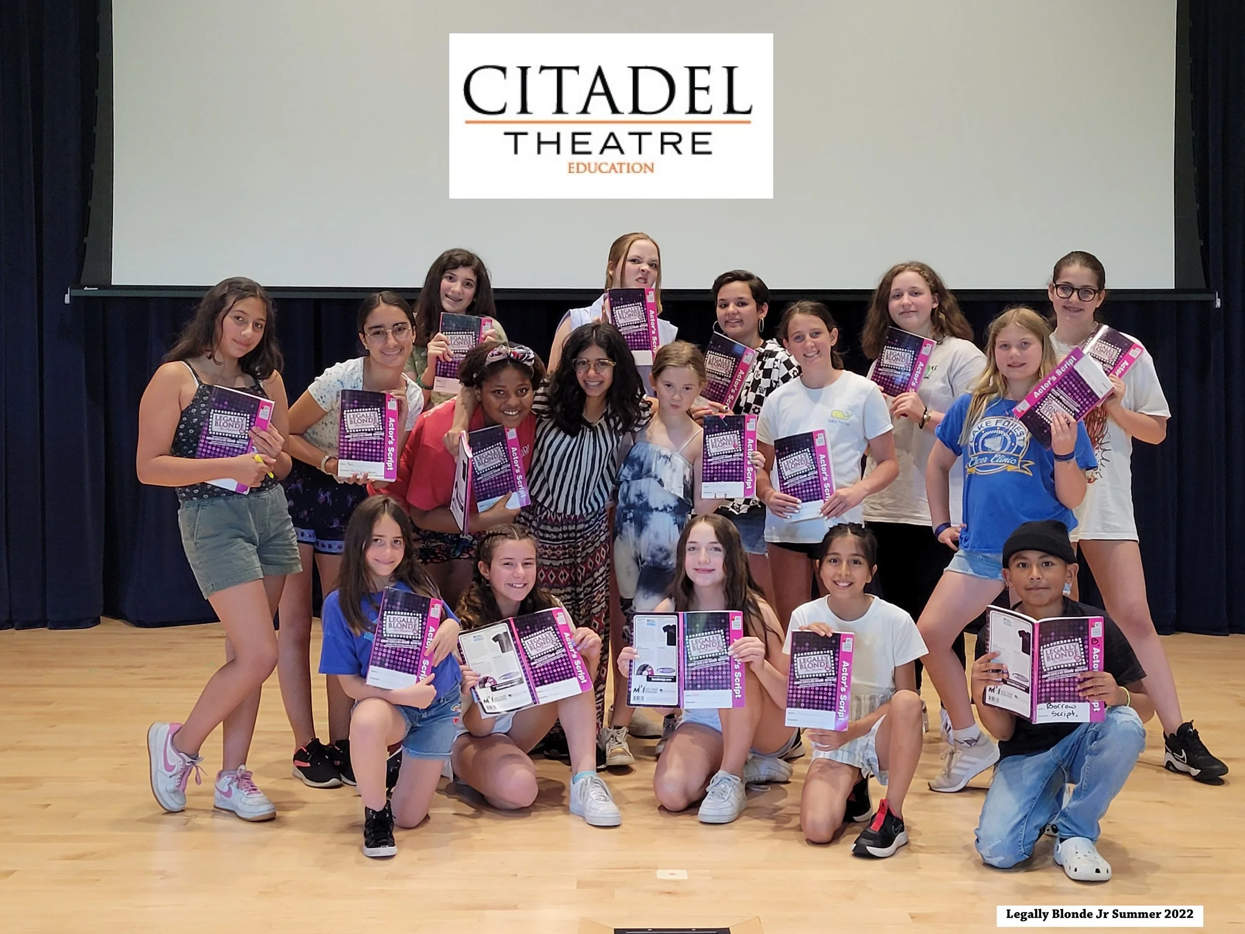 Group of children and a woman holding books on a stage with a large screen behind them displaying the Citadel Theatre Education logo. The children are smiling and posing for a photo.