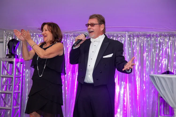 A man in a tuxedo singing into a microphone while a woman in a black dress claps and smiles on stage with purple and silver decorations in the background.