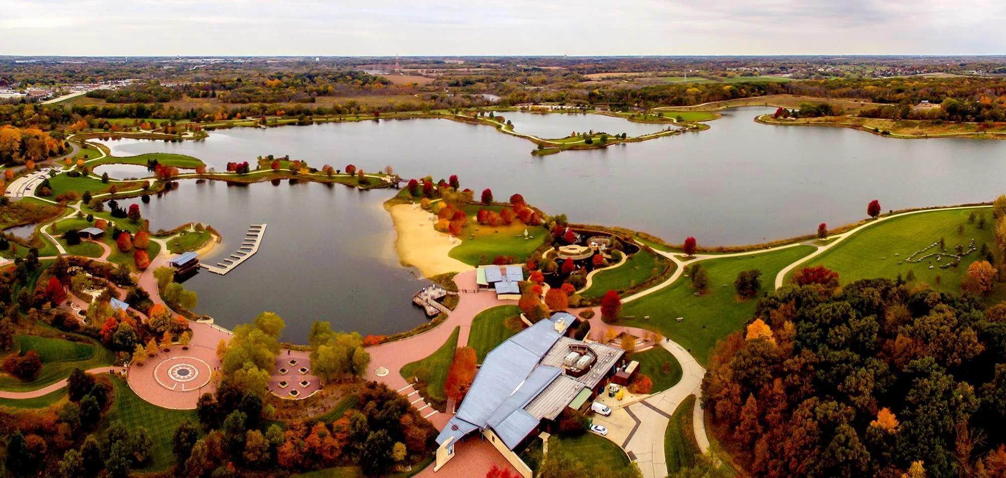 Aerial view of the Independence Grove Forest Preserve in Libertyville, IL with lakes, walking paths, trees with autumn foliage, and some buildings and parking areas.