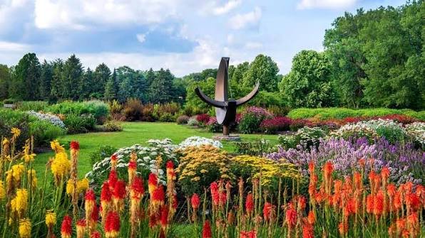 Colorful flower garden in the Chicago Botanic Garden with a statue in the center, surrounded by trees and a partly cloudy sky.