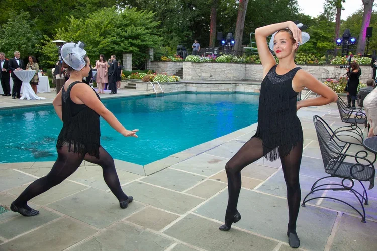 Two women dressed in flapper-style black dresses and headpieces performing jazz dance near a swimming pool at an outdoor event, with guests in formal attire and trees in the background.