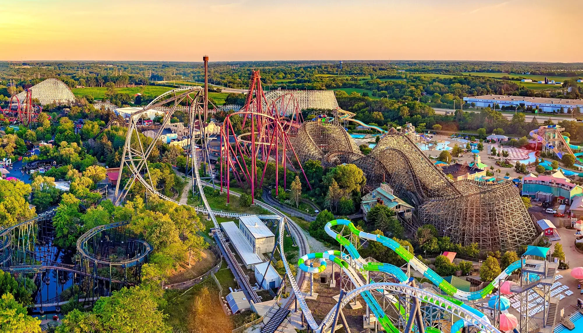 Aerial view of Six Flags Great America in Gurnee, IL with various roller coasters, water slides, and rides surrounded by green trees and open fields during sunset.
