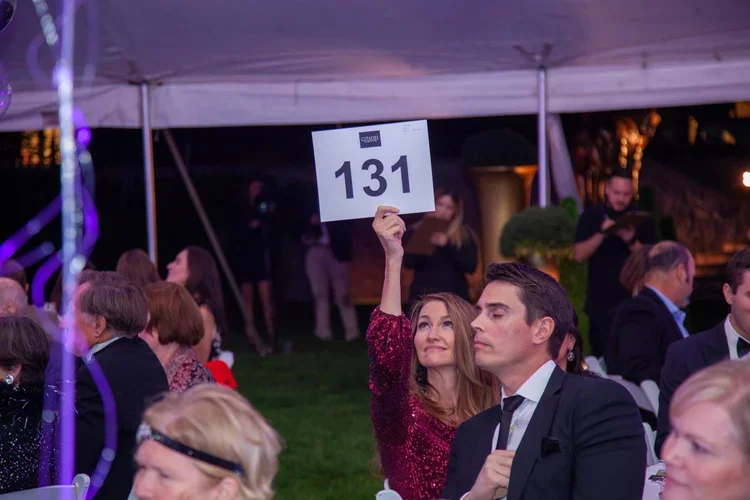 Woman in a red sparkly dress holding up a sign with the number 131 at a formal event seated at a table under a white tent.