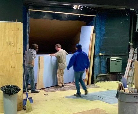 Three men install large sheets of drywall or similar material on a Citadel set, with construction tools and materials around them.
