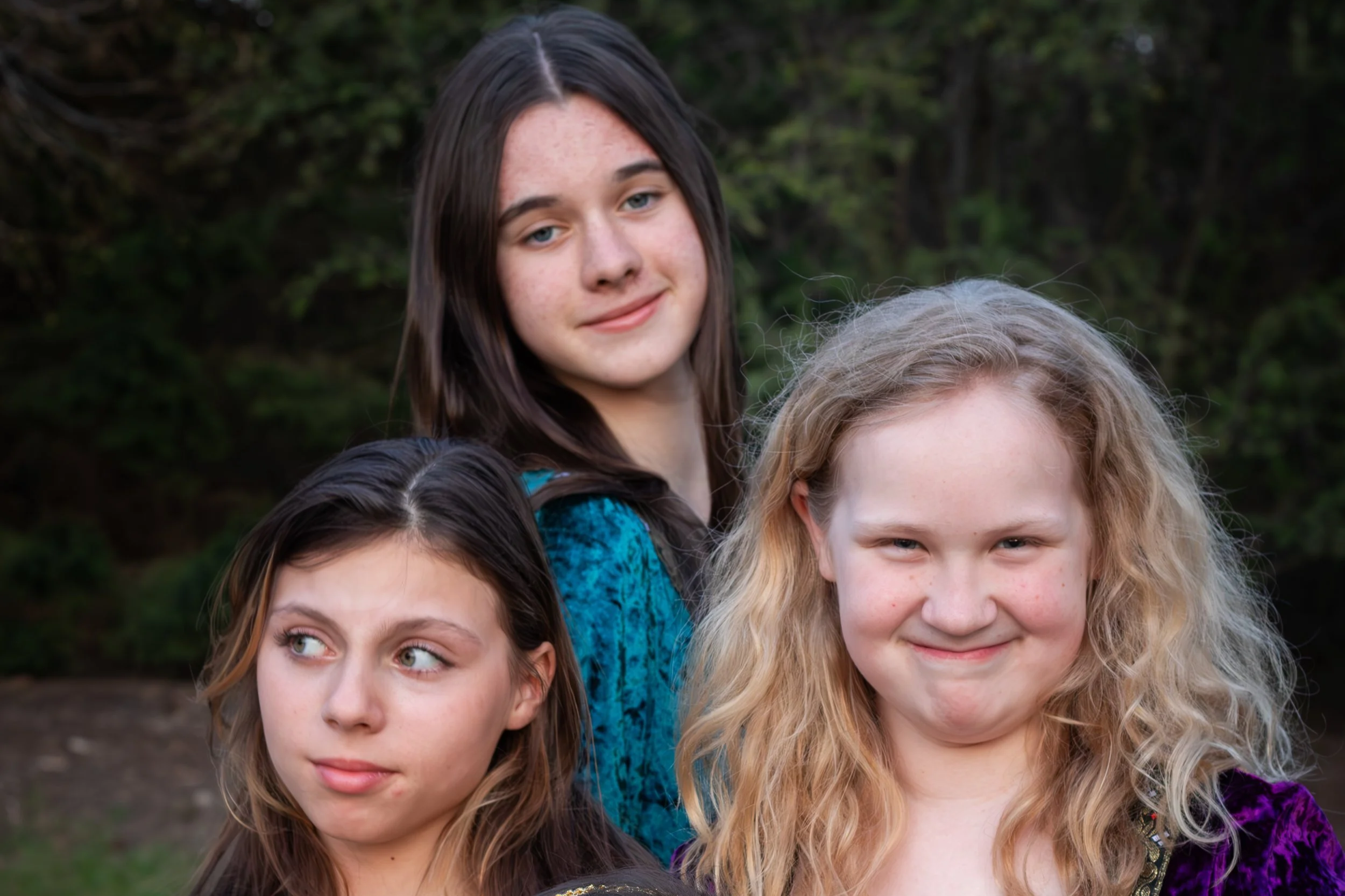 Close-up of a group of smiling girls with various hair colors and styles, outdoors with a wooded background.
