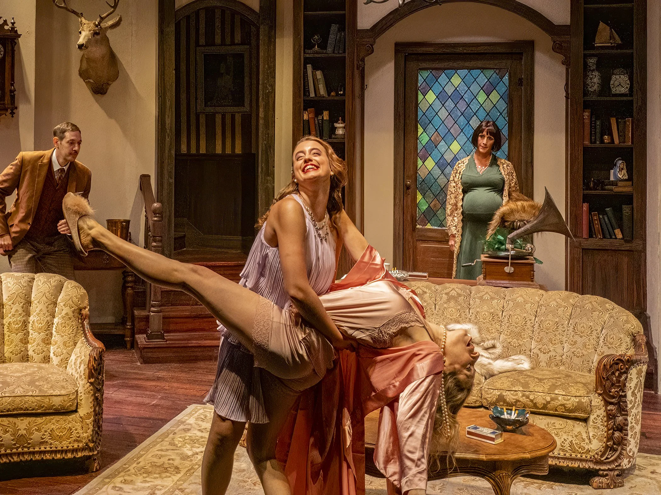 A scene from a Citadel's production of The Cottage set in a vintage-styled living room with a stained glass window. Two women are positioned in the foreground, dancing with two people observing in the background.