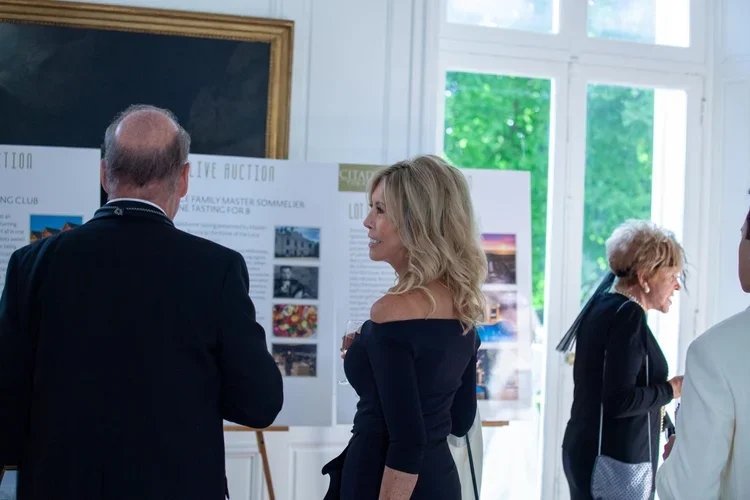 Three people in formal attire at an indoor event, looking at display boards with text and images, near large windows with green garden outside.