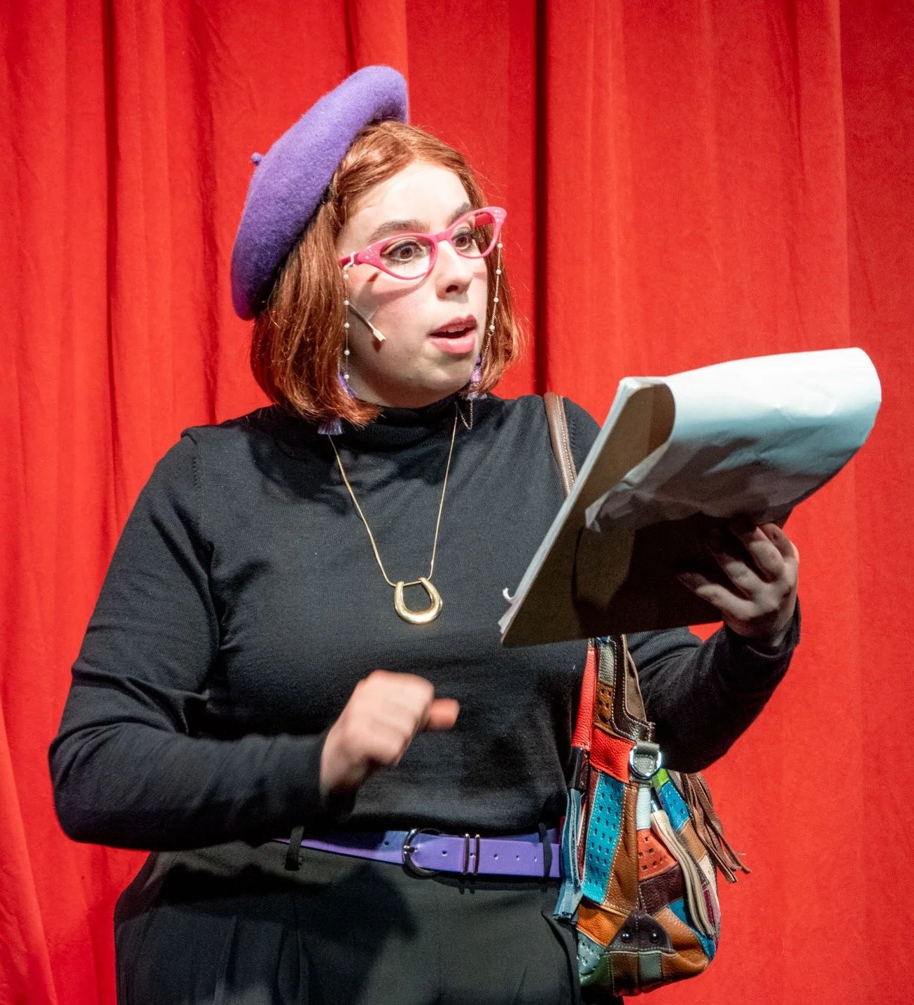 A woman with red hair, pink glasses, and a purple beret looks surprised while reading from a script in front of a red curtain.