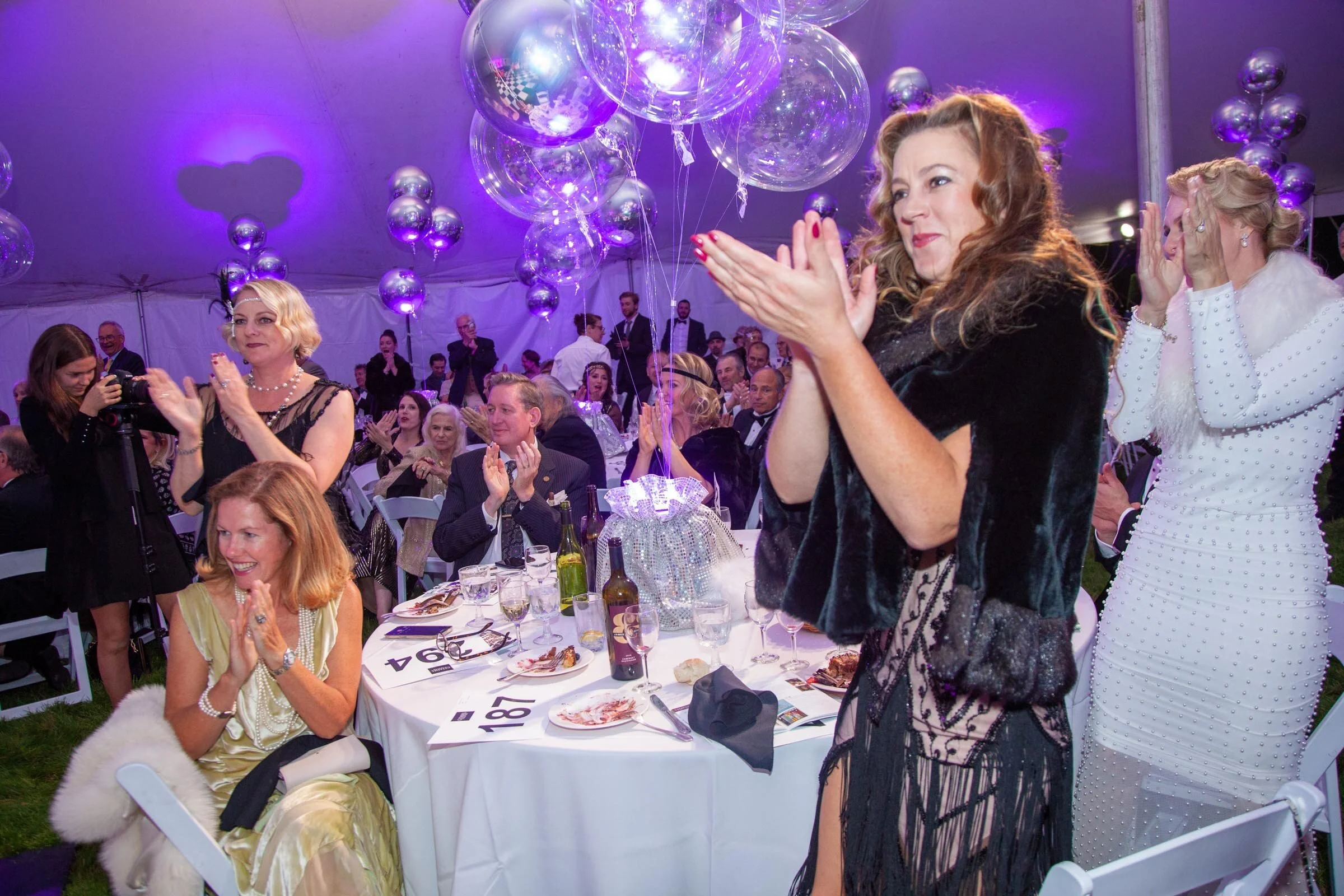 People gathered at Citadel's 12th Annual Gala inside a large tent decorated with purple and silver balloons, with some individuals clapping and others taking photos, enjoying a festive event.