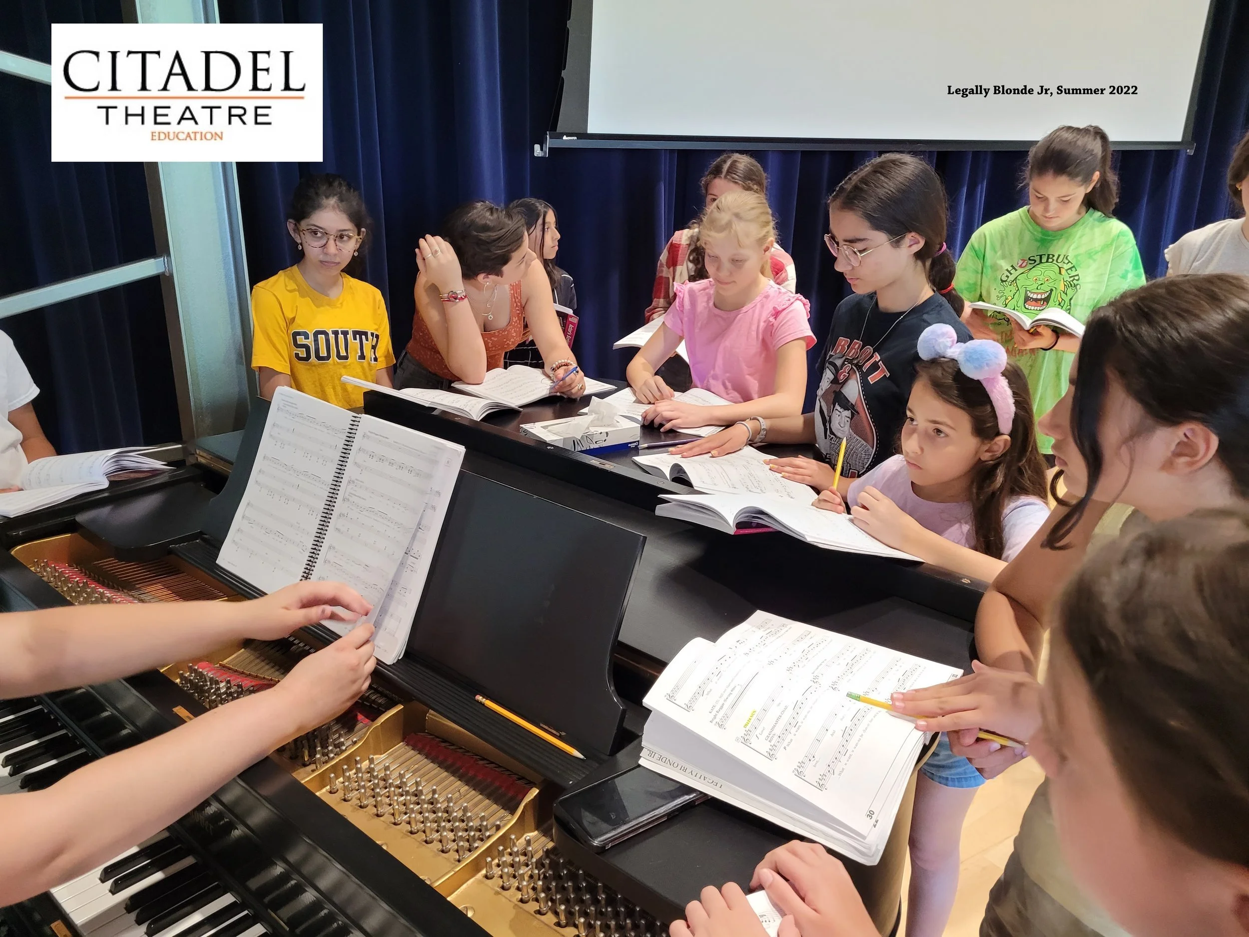 Group of children and adults gathered around a grand piano, reading sheet music, in a classroom or rehearsal space, with a music book on the piano.