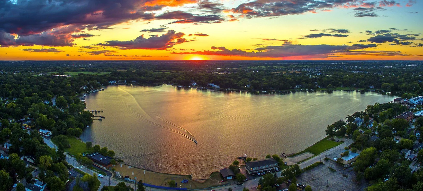 Aerial view of a large lake at sunset in Lake County, IL,, surrounded by green trees and residential houses, with a boat creating ripples on the water.