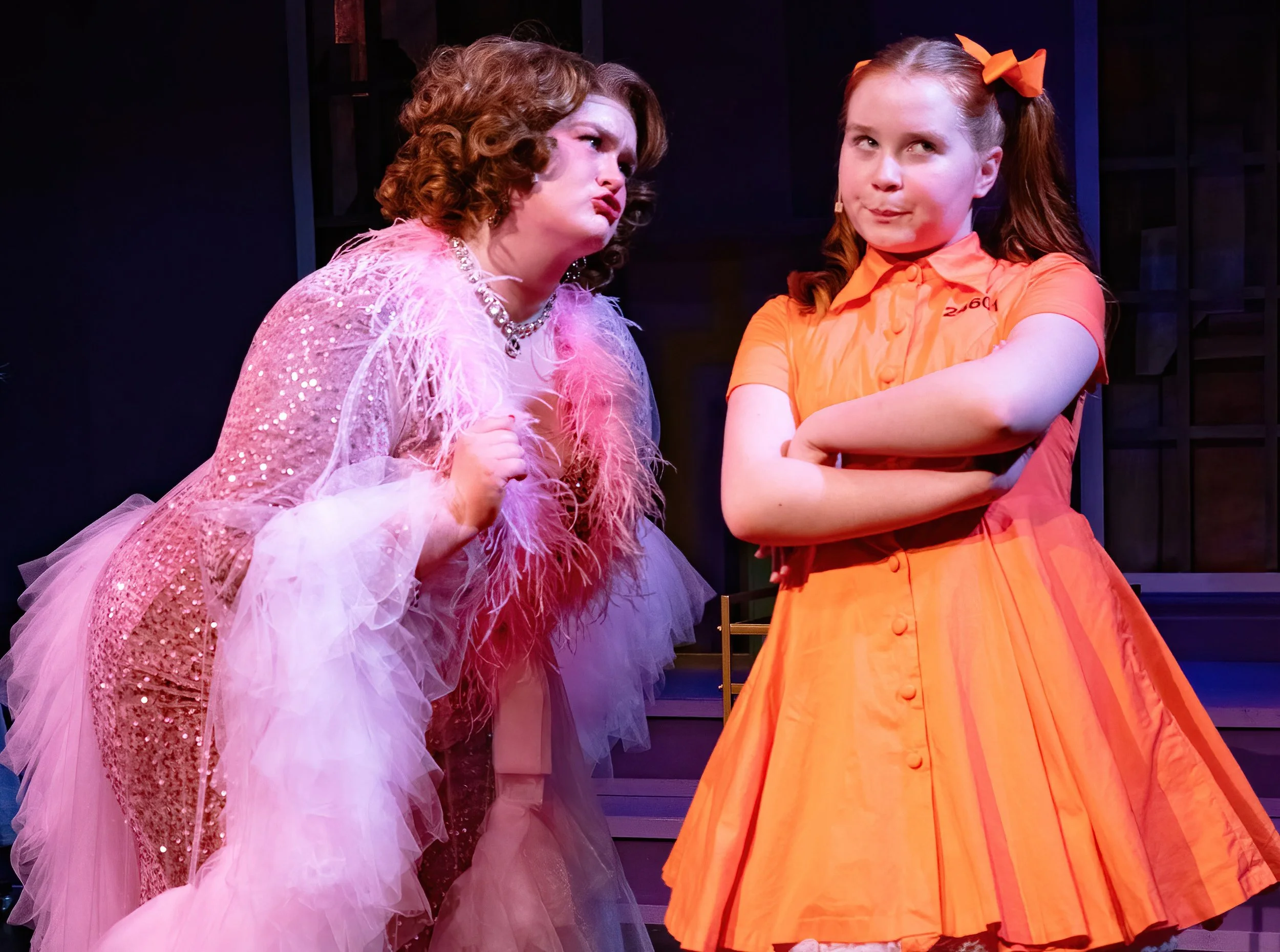 A woman and a girl in a performance of Ruthless at Citadel Theatre. The woman on the left is wearing a pink, sequined dress with feathers and has curly hair. The girl on the right, with hair in pigtails and orange bows wearing an orange dress