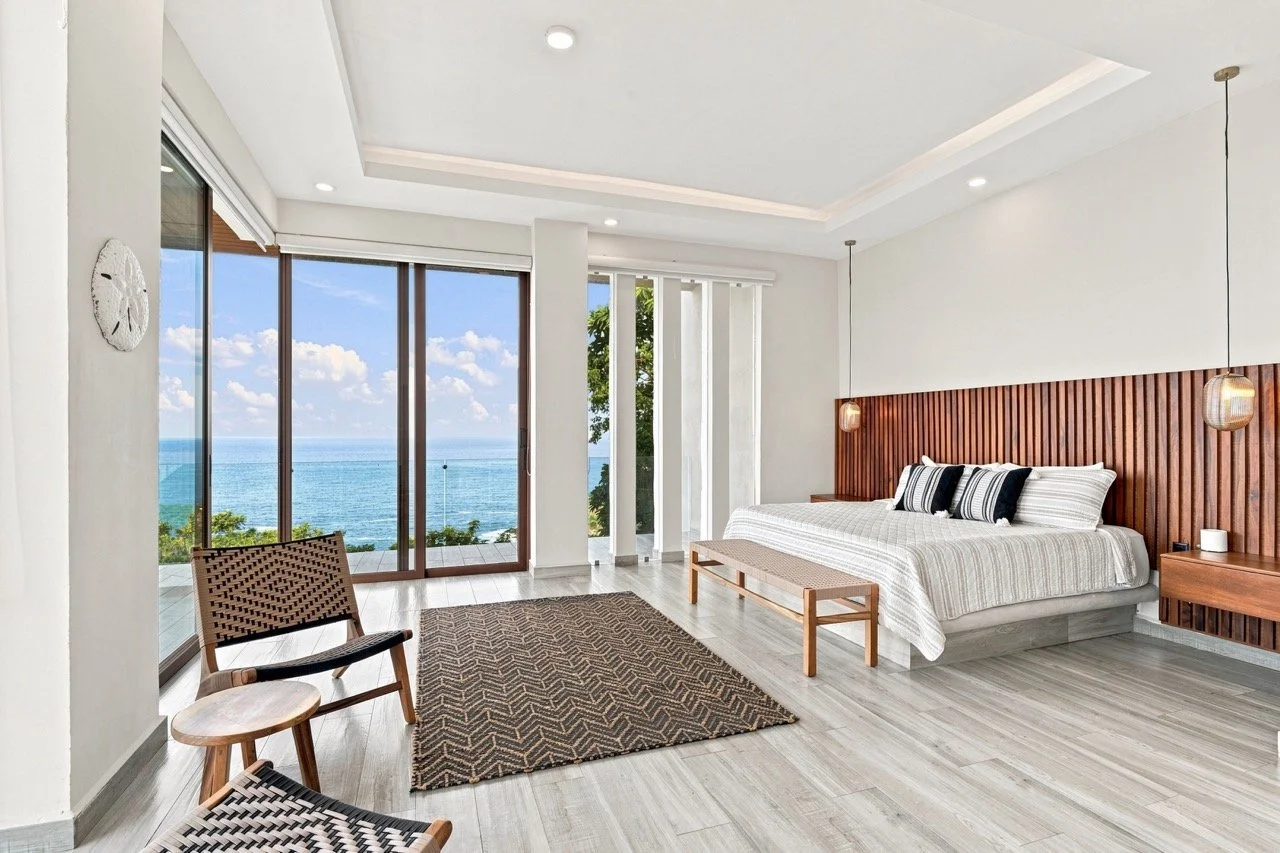 Habitación moderna con vista al mar, cama con cabecera de madera, ventanas panorámicas, suelo de madera claro, y decoración minimalista.
